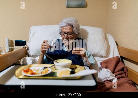 Una donna anziana siede comodamente nel suo letto con un vassoio di cibo e un bicchiere d'acqua, raffigurando un momento sereno in una struttura di assistenza che appe Foto Stock