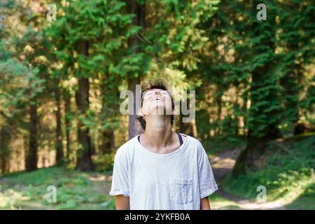 Un individuo non binario ride gioiosamente in una foresta illuminata dal sole, con gli occhi chiusi circondati da alti alberi verdi. Foto Stock