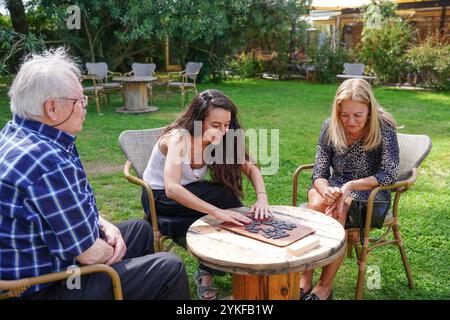 Una famiglia caucasica composta da due adulti anziani, una femmina e un maschio, e una femmina adulta più giovane, si impegna felicemente in un gioco da tavolo all'aperto surroun Foto Stock