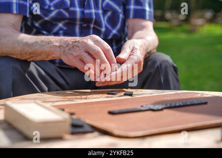 Le mani di un anziano uomo caucasico tagliato e irriconoscibile mentre gioca a un gioco da tavolo all'aperto, con pezzi di legno visibili su un tavolo illuminato dal sole. Foto Stock