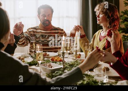 Famiglia caucasica che si tiene per mano e dice preghiera mentre si siede al tavolo delle feste durante le celebrazioni di Natale Foto Stock