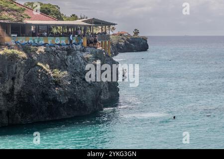 Un tuffatore da scogliera salta dalle rocce a Playa Forti a Westpunt, Curaçao, nel Mar dei Caraibi. Il ristorante Playa Forti è riconoscibile dalle lettere sulla parete rocciosa. Si trova direttamente sopra il punto di salto. I bagnanti stanno già nuotando nell'acqua sottostante. Foto Stock