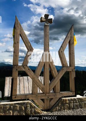 Un grande simbolo di legno con una croce si erge in modo prominente contro un drammatico cielo nuvoloso. Stemma dell'Ucraina. Situato in un'area montuosa, il Foto Stock