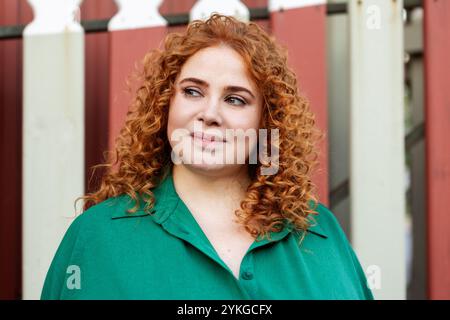 Ritratto di una bella donna Plus-size con capelli ricci rossi in un abbigliamento verde, che si affaccia sullo sfondo rosso all'aperto in estate Foto Stock