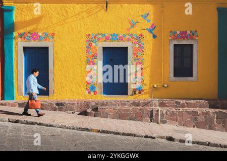 Guanajuato, Messico - 9 febbraio 2024: Un uomo cammina davanti a una vivace parete gialla adornata da un colorato murale floreale e colibrì, che mostra Guanaj Foto Stock