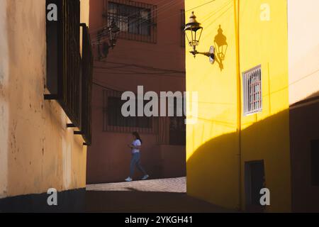 Guanajuato, Messico - 6 febbraio 2024: Una giovane donna del posto cammina attraverso un vivace vicolo illuminato dal sole a Guanajuato, Messico, con pareti gialle luminose circa Foto Stock