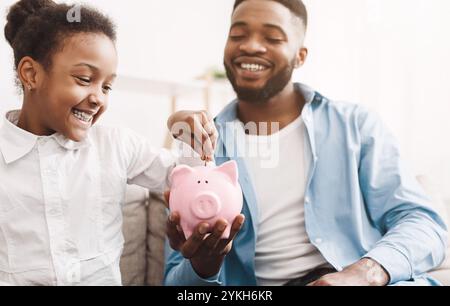 Risparmi personali. Piccola ragazza afro mettere il denaro a Piggybank Foto Stock