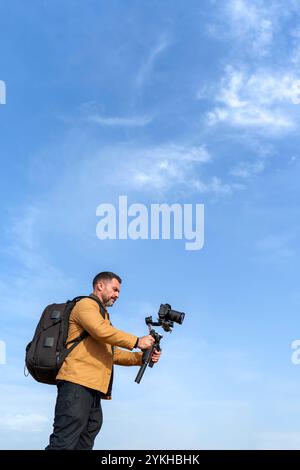 Operatore telecamera maturo che riprende con un Gimbal su uno sfondo blu chiaro Foto Stock