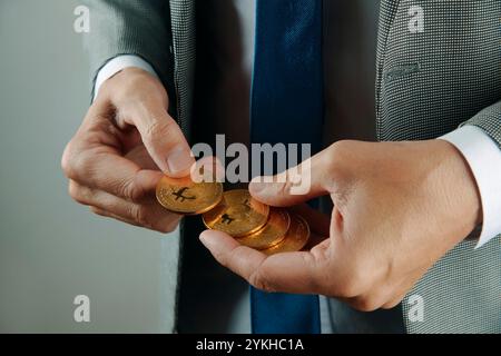 un uomo d'affari, che indossa un elegante abito grigio, una camicia bianca e una cravatta blu, ha dei bitcoin in mano al chiuso Foto Stock