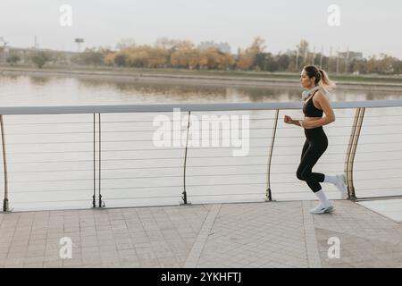 Un determinato corridore procede con sicurezza lungo un sentiero lungo il fiume, abbracciando l'aria frizzante del mattino, con lo skyline della città e gli alberi autunnali che offrono un'immagine Foto Stock