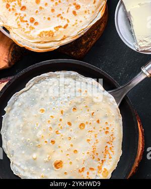 Friggere crepe sottili in una padella, sullo sfondo con crepe pronte e burro per loro, Foto Stock