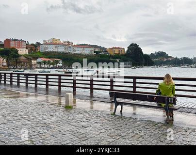 Vista posteriore di una donna seduta sulla panchina che guarda le barche nel porto di Mugardos, Galizia, Spagna Foto Stock