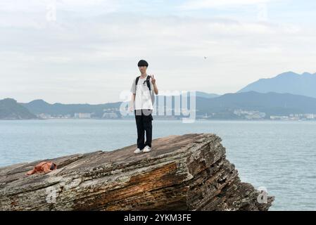 Un turista che posa dalla cima una grande roccia sulla costa meridionale dell'isola di Tung Ping Chau a Hong Kong. Nan'Ao Town, nella provincia del Guangdong, sul retro. Foto Stock