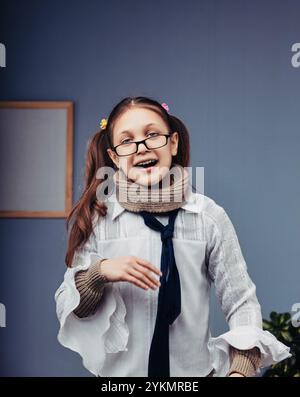 Ritratto di una allegra ragazza dell'età elementare che indossa occhiali da vista, una sciarpa e una camicia bianca, sorridente e guarda la fotocamera Foto Stock