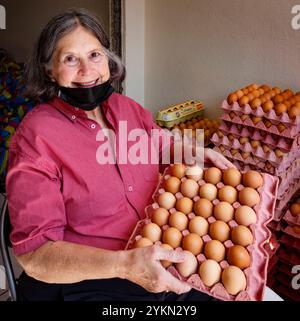 Sorridente anziana signora tiene un vassoio di uova marroni in un mercato locale, indossando una maschera facciale Foto Stock