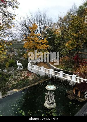 Tranquillo giardino caratterizzato da una statua di cervo che si affaccia su una fontana e uno stagno, circondato da un vivace fogliame autunnale in un ambiente sereno. Kamyanets Pod Foto Stock