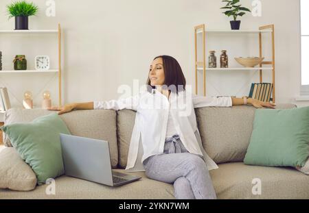 Donna adulta sorridente e calma seduta sul divano vicino al computer portatile, che si rilassa a casa. Foto Stock