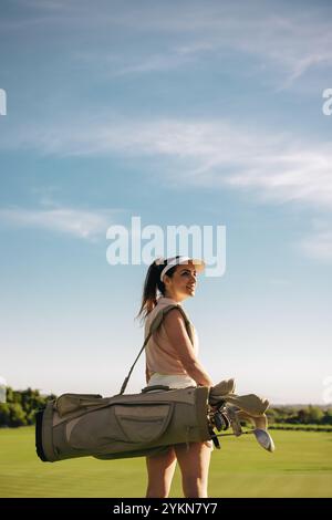 Golfista sorridente che porta mazze da golf con abbigliamento da golf in piena luce del sole, pronta per una partita di golf in una bella giornata. Foto Stock