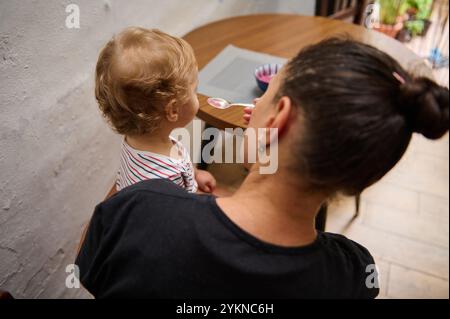 Una madre nutre teneramente il suo bambino in una cucina calda e accogliente. Il bambino, seduto in grembo, anticipa il prossimo cucchiaio. L'impostazione riflette un'alimentazione Foto Stock