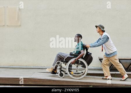 Foto a tutta lunghezza minima di un uomo afro-americano con disabilità su sedia a rotelle e salendo sulla rampa con un amico che aiuta a copiare lo spazio Foto Stock