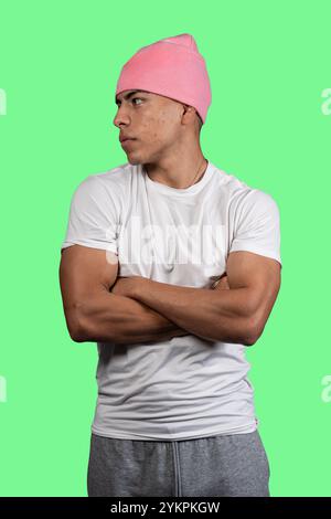 Foto in studio di un giovane latino che indossa un berretto rosa, una t-shirt bianca e pantaloni in felpa grigi, che incrociano le braccia e guardano lateralmente su un vivace gr Foto Stock