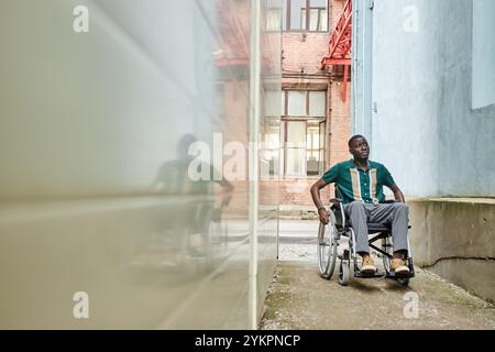 Riprese grandangolari di un uomo afroamericano con disabilità che utilizza la sedia a rotelle in città e si sposta verso la fotocamera su un marciapiede stretto tra gli edifici e lo spazio di copia Foto Stock