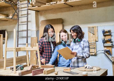 Un gruppo di tre donne che collaborano in un bosco. Sono impegnati in un apprendistato, discutendo di piani mentre sono circondati da strumenti per la lavorazione del legno. Foto Stock