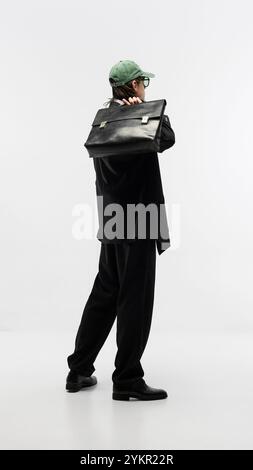 Immagine a figura intera di un giovane uomo che indossa un costume total black, un berretto da baseball verde e una valigetta in pelle, che posa di nuovo uno sfondo bianco dello studio Foto Stock