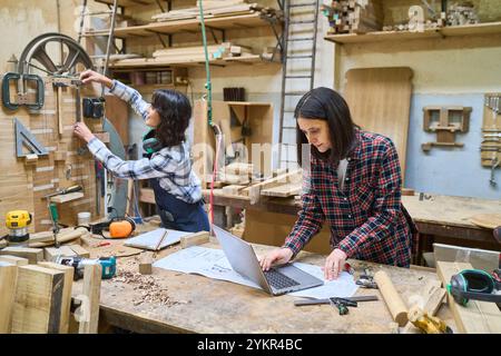 Due donne che lavorano insieme in un bosco, concentrandosi sul lavoro di squadra e sullo sviluppo delle competenze. Sono impegnati in attività di lavorazione del legno circondati da utensili e. Foto Stock