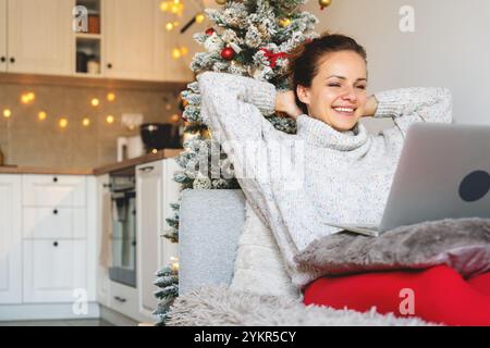 Durante le vacanze di Natale, la donna si siede comodamente sul divano e utilizza il proprio computer portatile. Foto Stock