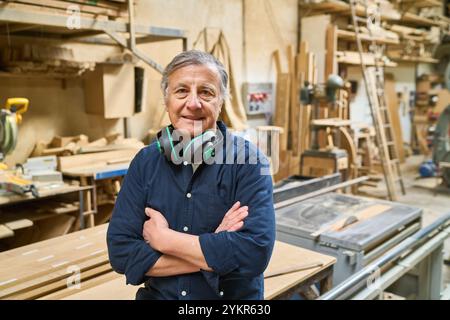 Artigiano esperto posa con fiducia in un vivace laboratorio, simboleggiando la dedizione e l'esperienza nella lavorazione del legno. Circondato da strumenti e materiali Foto Stock