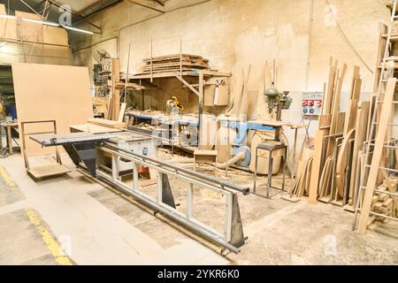 Vista interna di un'officina di legname con varie attrezzature per la lavorazione del legno, legno impilato e attrezzi. Lo spazio organizzato mette in risalto il mach industriale Foto Stock