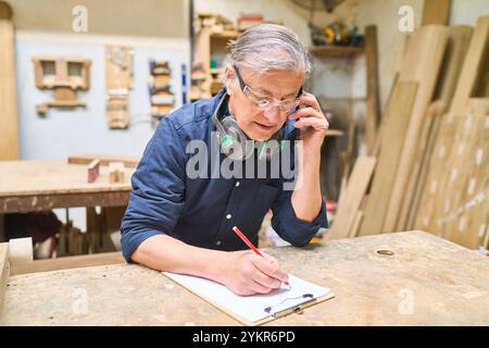 Lavoratore anziano in un'area boschiva che gestisce le attività mentre parla al telefono. Dimostra organizzazione e comunicazione in un ambiente di lavorazione del legno. Foto Stock