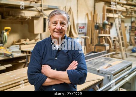 Un artigiano anziano si trova in un bosco circondato da utensili e materiali per la lavorazione del legno, mostrando anni di esperienza nella falegnameria e nel Foto Stock