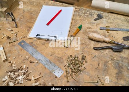 Utensili e materiali assortiti per la lavorazione del legno sono sparsi su un banco da lavoro rustico. La scena include strumenti come un righello, un cacciavite e un pennello, enfatizza Foto Stock