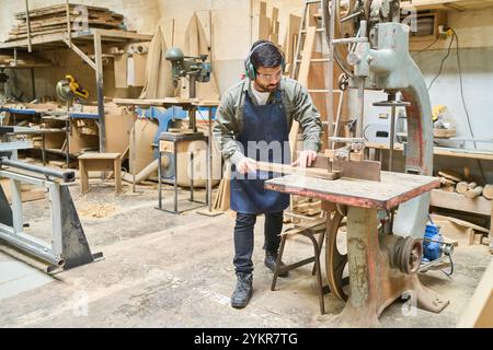 Un carpentiere esperto che indossa dispositivi di protezione aziona una sega per tagliare una tavola di legno in un'officina. L'area di lavoro è piena di utensili per la lavorazione del legno e tappetino Foto Stock