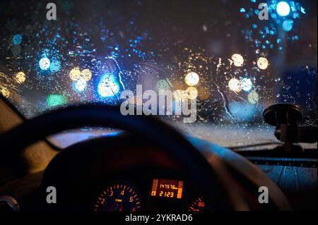 Gocce di pioggia sul parabrezza di un'auto con luci urbane sfocate e colorate di notte, viste dall'interno. Volante e cruscotto illuminati con luce soffusa Foto Stock