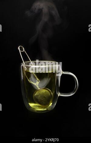 Tazza in vetro trasparente doppia, con tè verde e setaccio per tè con morsetto in argento, vapore visibile, isolata sul nero Foto Stock