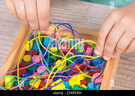 Mani del bambino che giocano a filo e perline colorate in legno utilizzate per realizzare braccialetti. Sviluppo delle capacità motorie dei bambini, coordinamento, creatività e logica Foto Stock