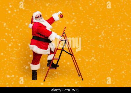 Completa il profilo di dimensioni foto a lato di funky divertente babbo natale decorare albero di natale salire le scale desidera appendere X-mas ornamento sulla elevata usura superiore rosso rosso alla moda Foto Stock