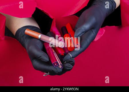 Mani con i guanti che tengono in mano vari bastoncini lucidi per labbra. Sfondando un muro di carta rosso Foto Stock