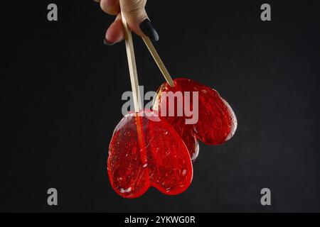 Mano che regge caramelle rosse a cuore su sfondo nero. Un paio di dolci lecca lecca-lecca Foto Stock