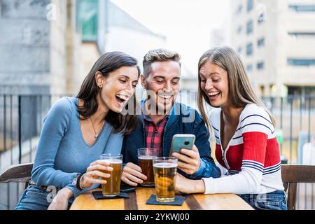 Tre amici che ridono insieme mentre guardano uno smartphone all'aperto. Giovani adulti che si godono un momento di divertimento con una birra in un bar della città Foto Stock