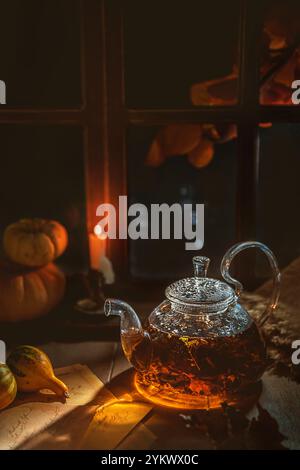 Teiera di vetro con tè in fiore, candela in fiamme, zucche e lettere vicino alla finestra in una stanza buia che crea un'atmosfera accogliente in autunno Foto Stock