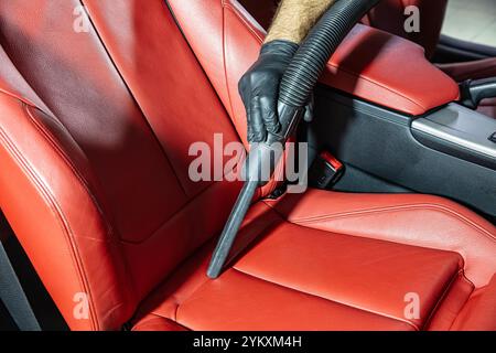 Un uomo che indossa guanti aspira un'auto con interni in pelle. Una panoramica dettagliata dell'aspirazione di un seggiolino in pelle rossa per garantire una pulizia accurata di Foto Stock