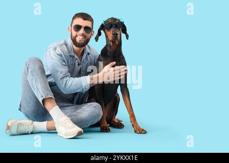 Giovane con il cane Dobermann in occhiali da sole seduto su sfondo blu Foto Stock