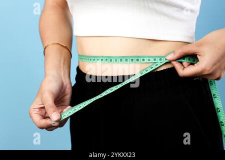 Slim Asian Young Woman che misura la sua vita usando il nastro di misurazione, isolato su sfondo blu Foto Stock