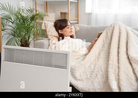 Giovane donna con una coperta calda che legge un libro sul divano vicino al riscaldatore elettrico a convettore a casa Foto Stock