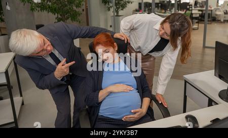 Un anziano uomo e una donna caucasica guardano disapprovatamente al loro collega incinta che dorme in ufficio. Foto Stock