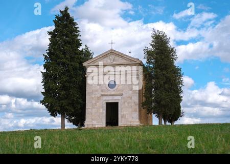 Incastonata tra le dolci colline della Toscana, una pittoresca cappella sorge senza tempo, incorniciata da alti cipressi e da un cielo azzurro vivido, invitando i visitatori ad uscire Foto Stock